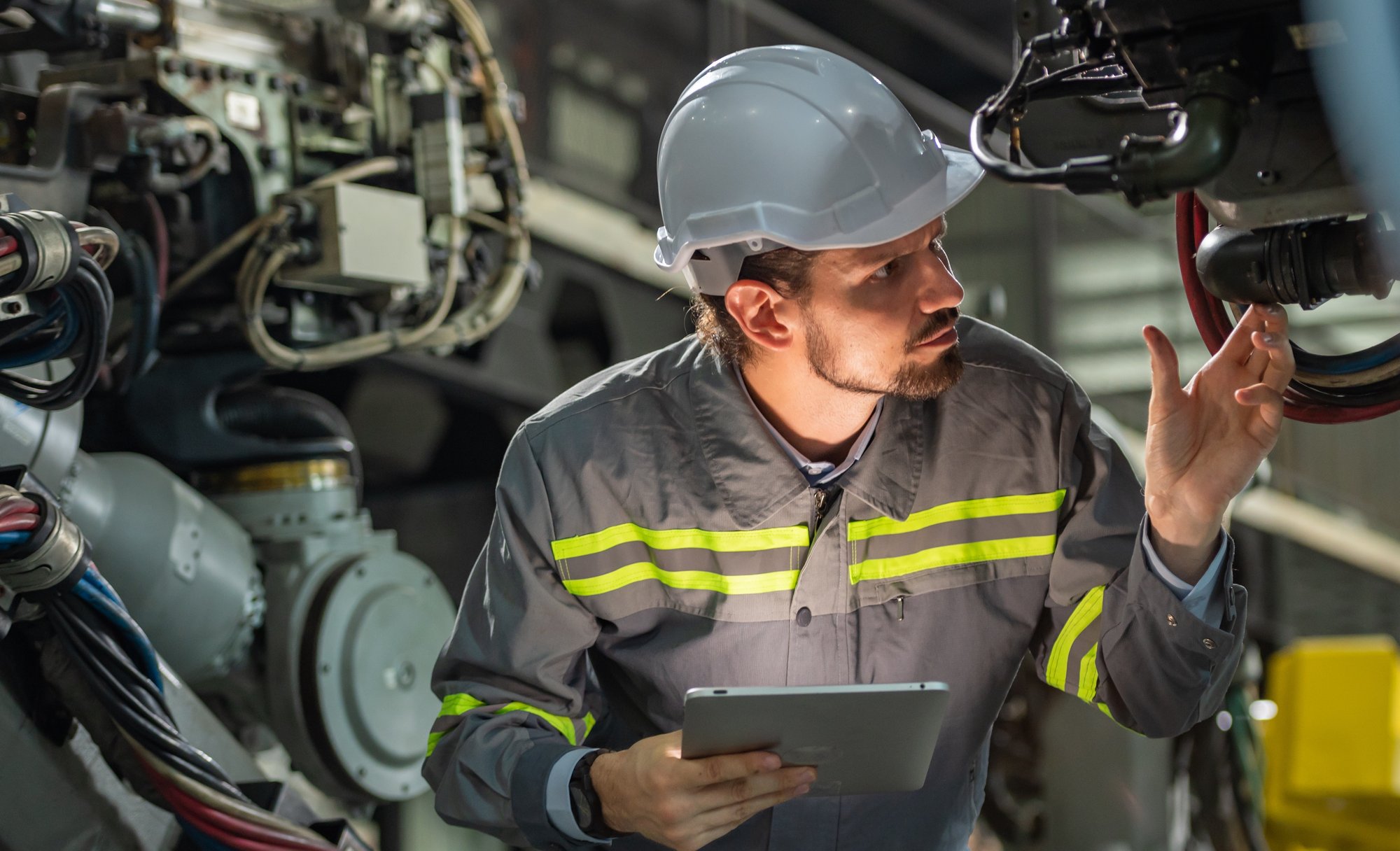 A man with a tablet in hand, wearing a helmet and safety clothing, is looking at the electrical system of a company.