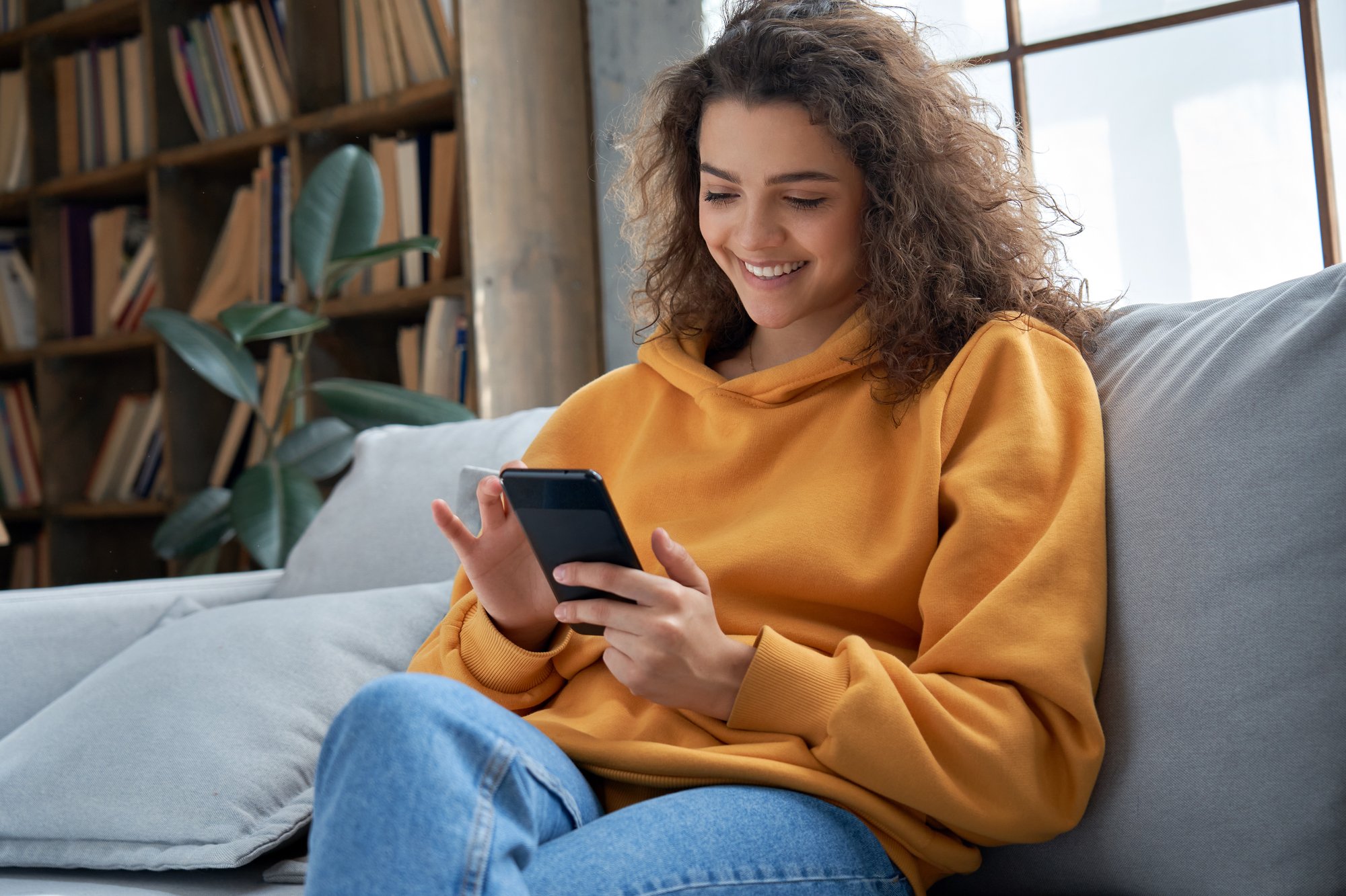 A smiling girl in a yellow hoodie is sitting on a couch, holding a smartphone.