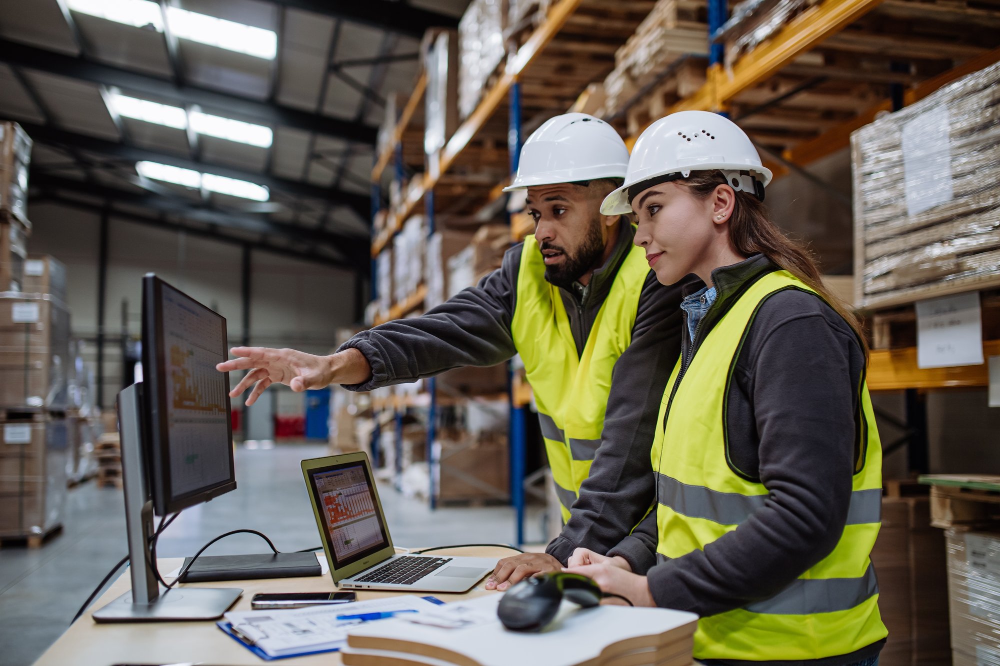 A man and two women are in a warehouse of an Industry 4.0 setting, discussing while looking at a tablet.