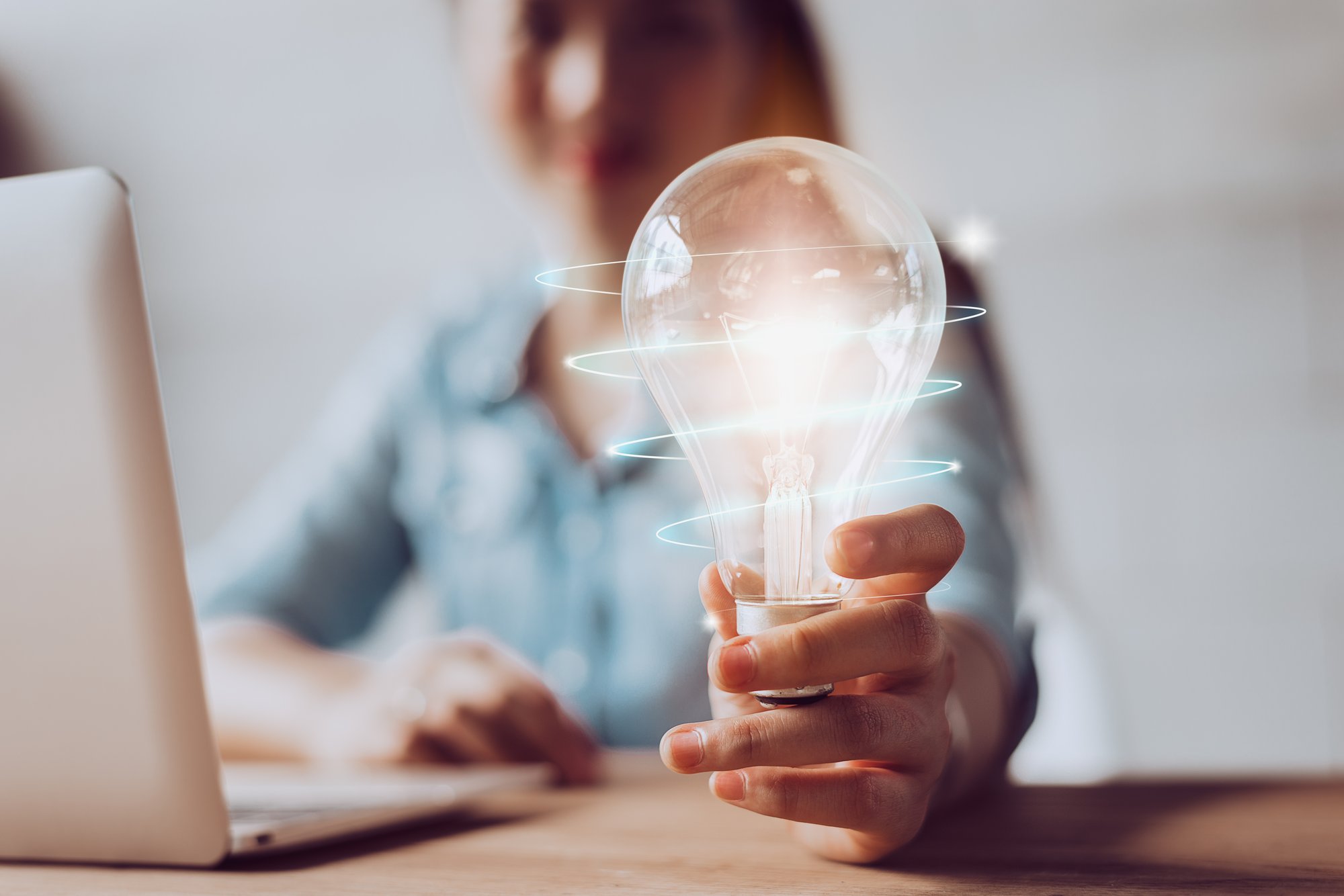 In the blurred background, a person is sitting in front of a computer, holding a lit lightbulb surrounded by a neon glow.