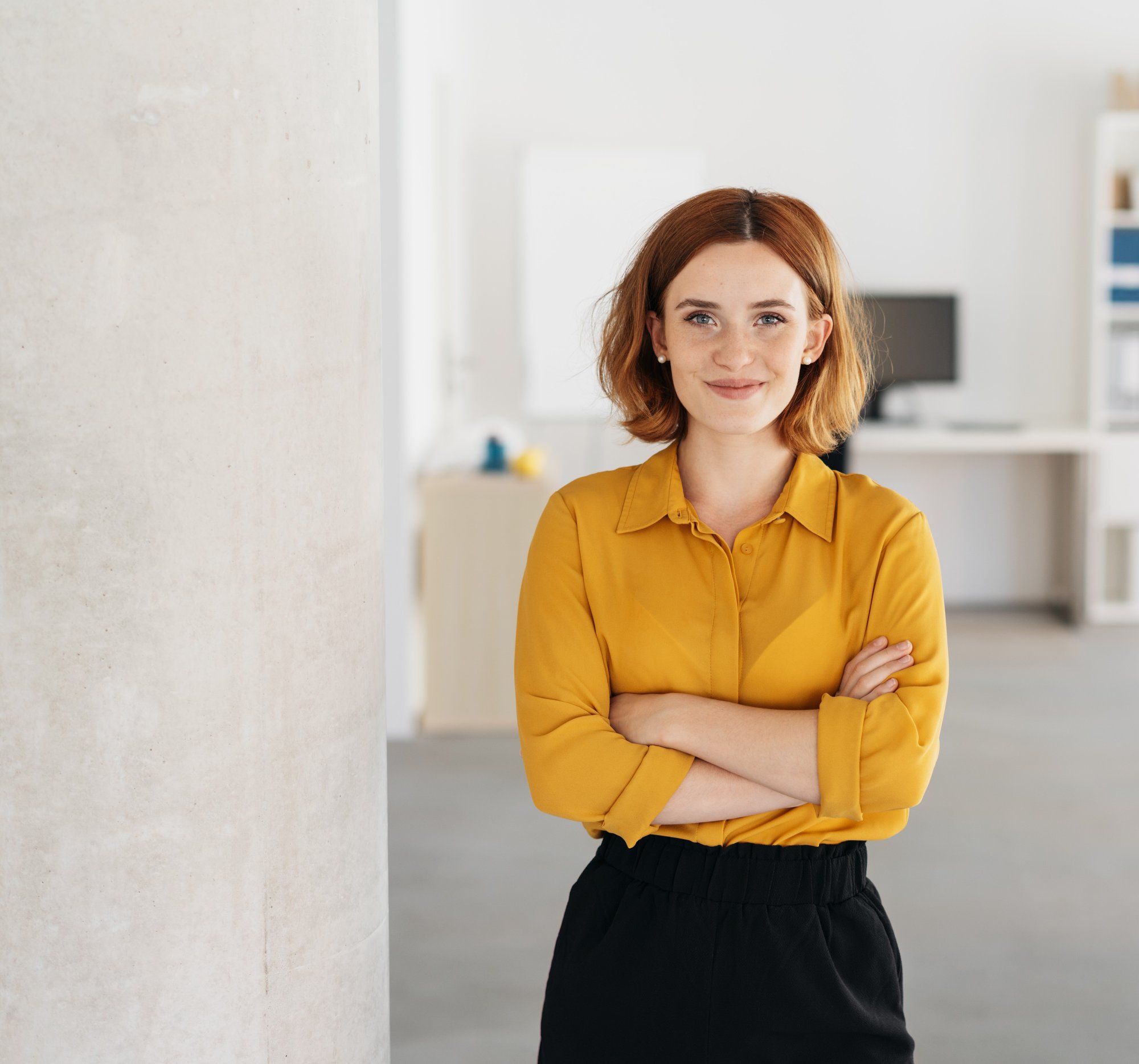 A young, happy, relaxed, and self-assured businesswoman standing with her arms crossed in a spacious office. 