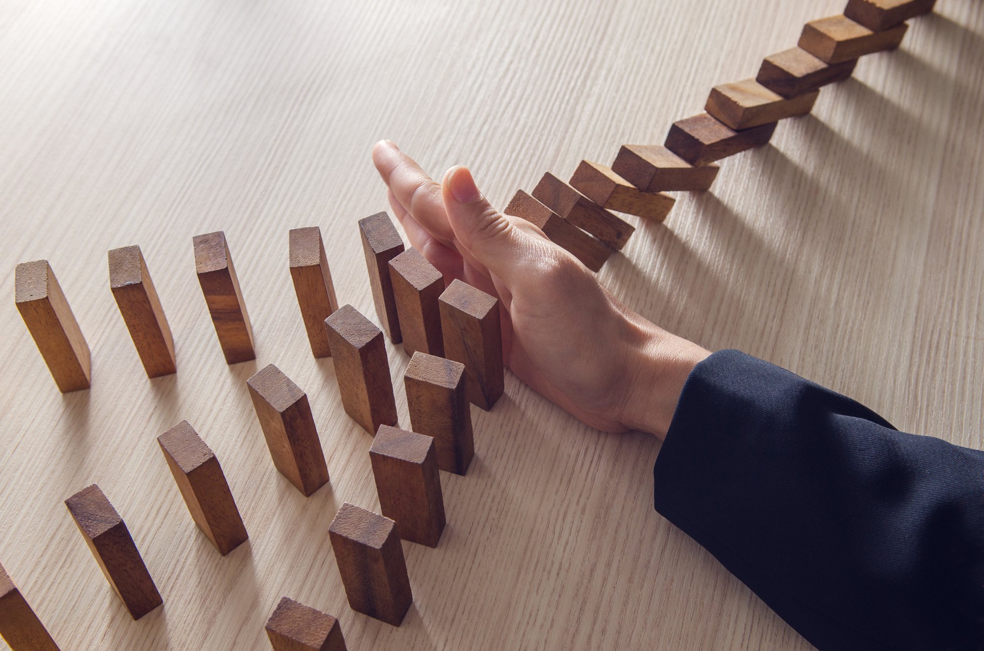 Primo piano di una mano di un uomo che stoppa l'effetto domino di alcuni mattoncini in legno messi in fila uno dietro l'altro. 